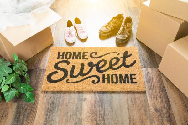 Home Sweet Home Welcome Mat, Moving Boxes, Women and Male Shoes and Plant on Hard Wood Floors. Home Sweet Home Welcome Mat, Moving Boxes, Women and Male Shoes and Plant on Hard Wood Floors.